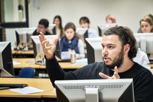 A student is talking and gesticulating in a classroom with computers and students. Photo by Håkan Röjder.