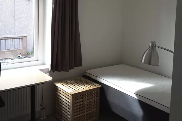 Overview of room C1006 at housing area Eddan showing the desk by the window, the laundry basket and the bed to the right.