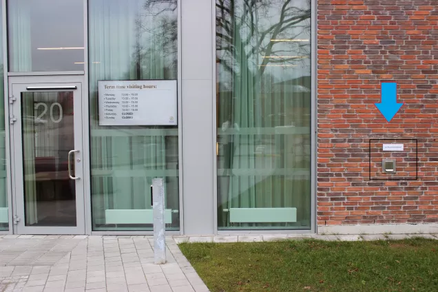 A glass reception entrance in a red brick building at Tunavägen 20, with an added blue arrow pointing out a key return chute to the right of the entrance. Edited photo by LU Accommodation.