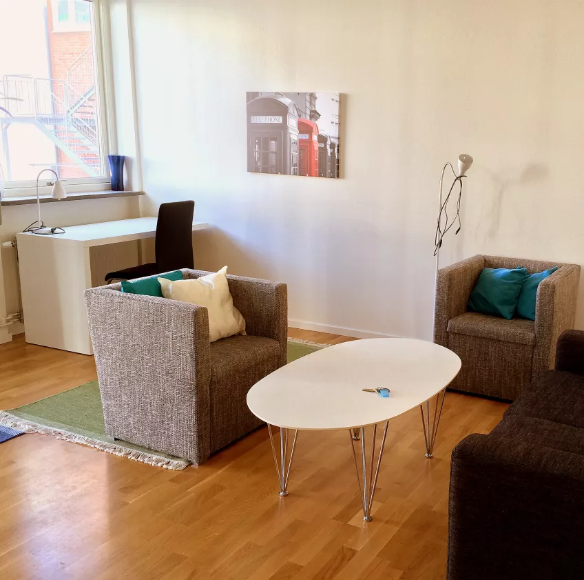 A living room witha sofa and two armchairs around a coffee table and a desk in the background at the housing area Folkets park (picture).