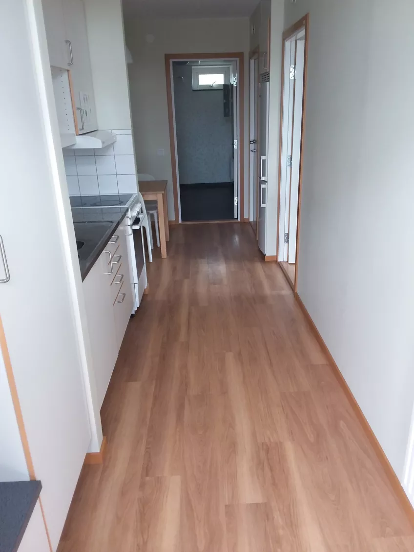 Kitchenette in apartment 6V at the housing area Bautastenen showing the sink, cooker and kitchen table to the left, the open bathroom door in the middle and the doors to the separate room and refrigerator/freezer to the right (picture)