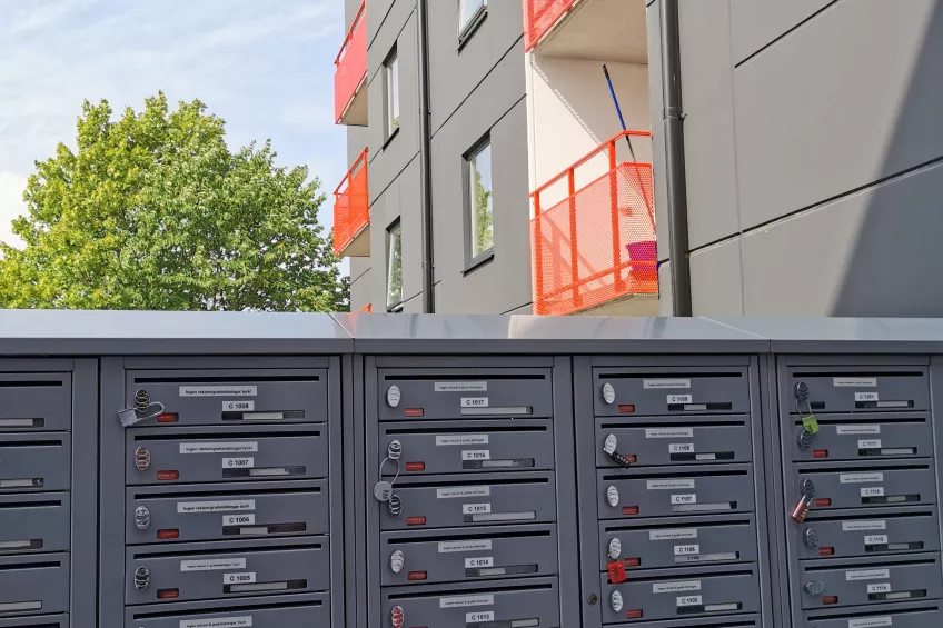 At the bottom letter boxes in dark grey and beyond them parts of the housing area Eddan showing to the upper right in grey, white and red, to the upper left blue sky and the top of a green tree (picture)